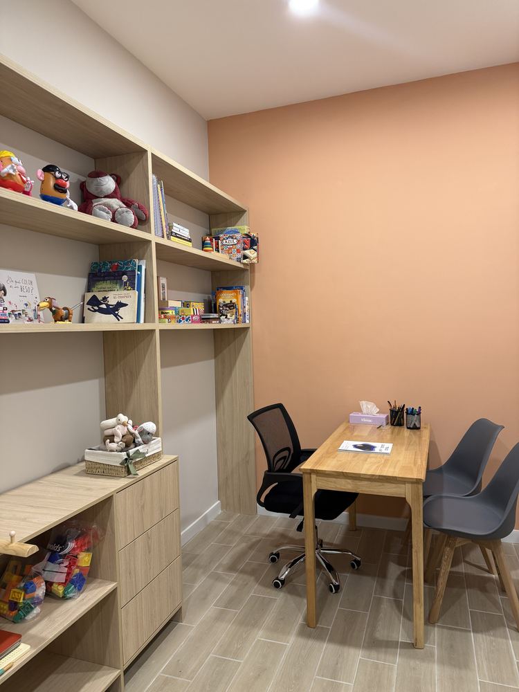 Consultorio de psicología: librero con cuentos y juegos, escritorio de madera y muro durazno.